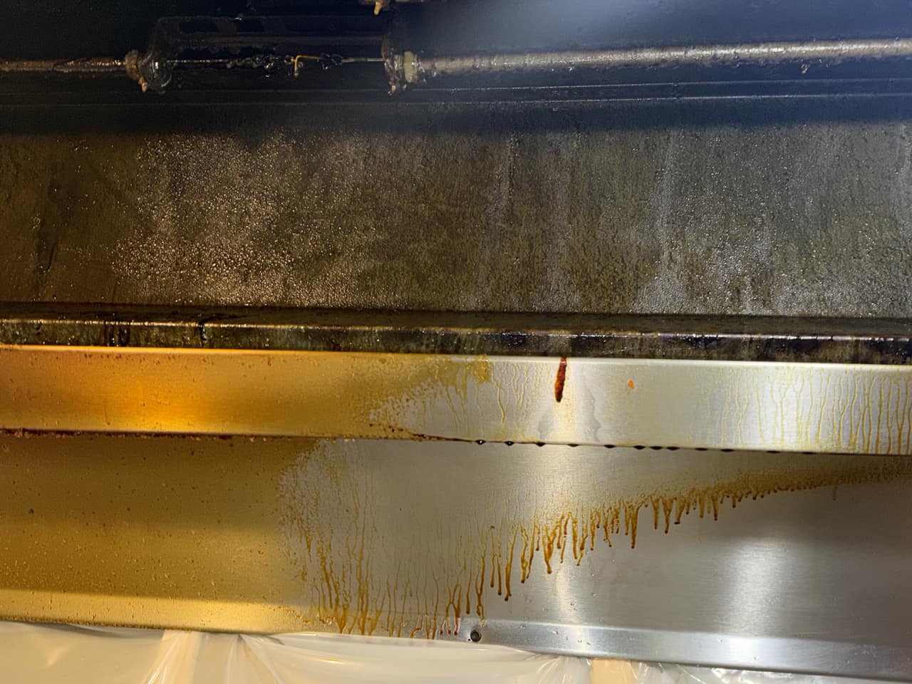 Inside a barbecue kitchen's exhaust hood before cleaning: the plenum wall is coated in grease and brown grease runs drip down the stainless-steel lip below it.